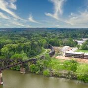 A river flows under a railway bridge through a lush green forest, buildings and a blue sky overhead.