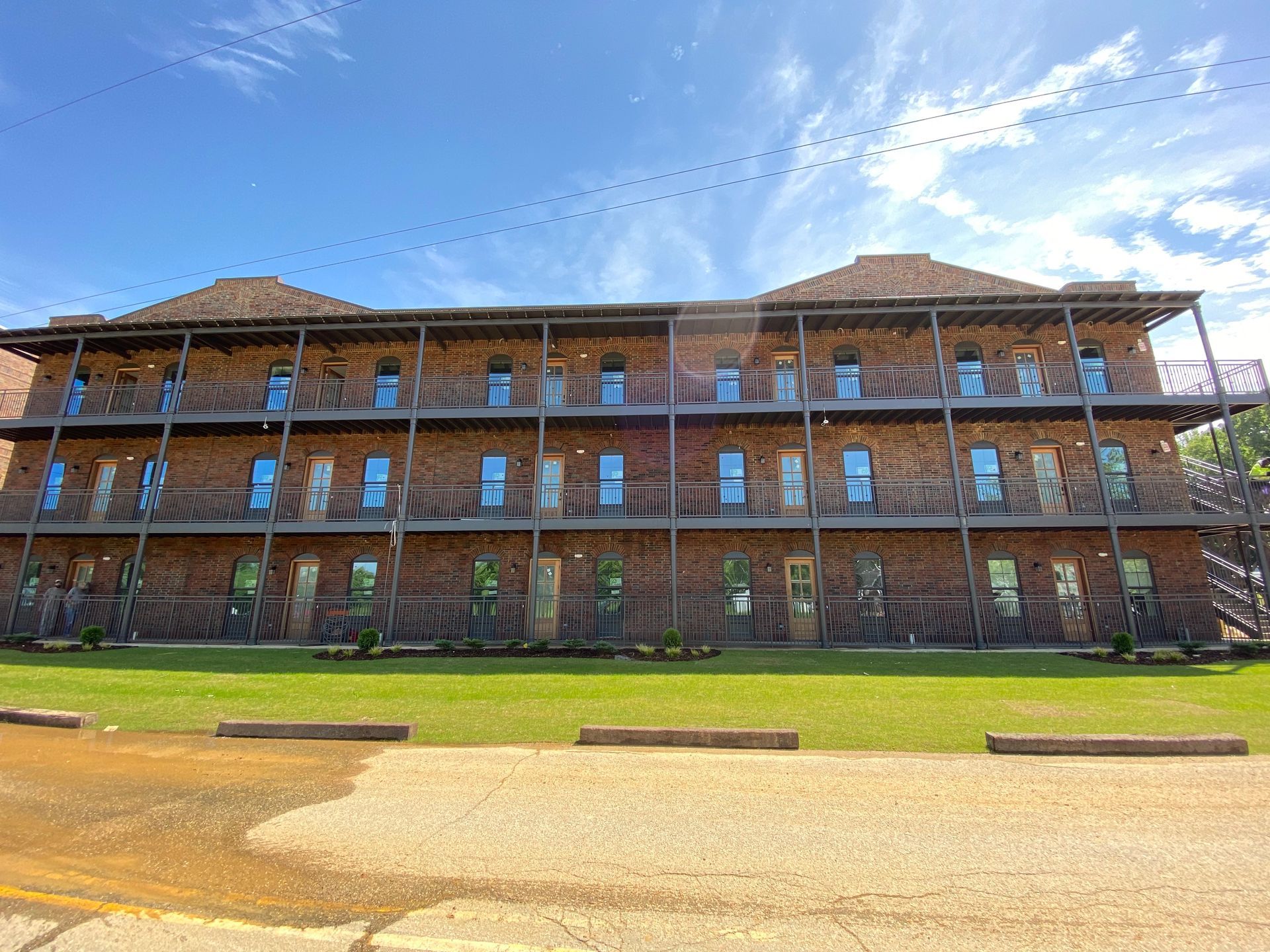 Three-story brick building with balconies and many windows, under a blue sky, on a grassy lot.