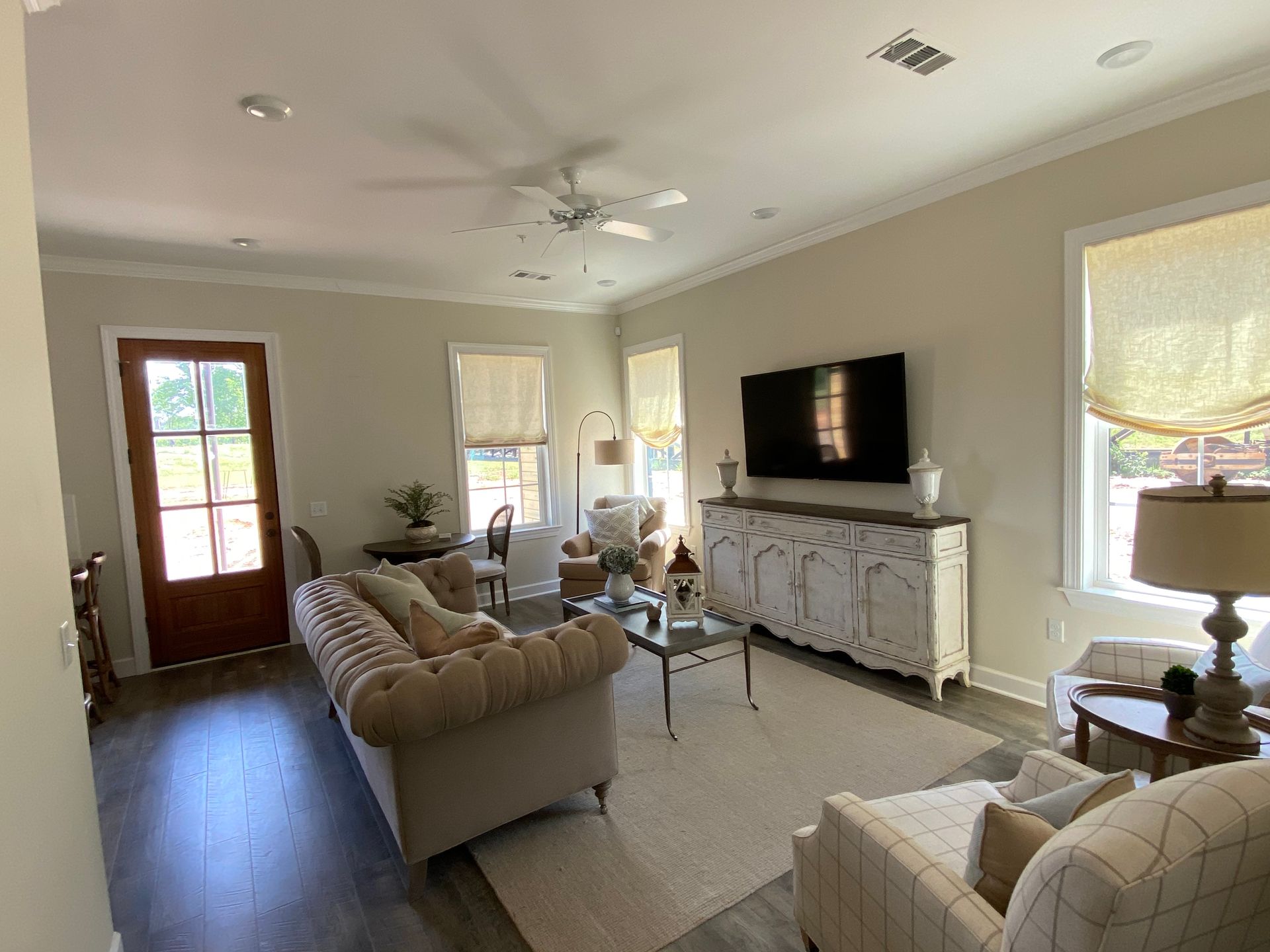 Living room with beige walls, wood floor, white trim. Furniture includes sofa, chairs, TV, and table. Natural light from windows.