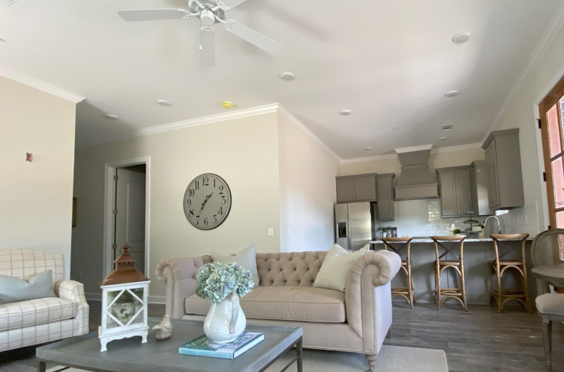 Living room with beige sofa, grey kitchen cabinets, large clock, and natural light.