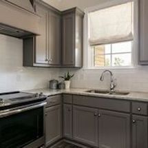 Gray kitchen cabinets with stainless steel appliances, white backsplash and roman shade.
