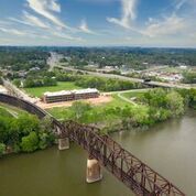 Aerial view of a brown metal bridge over a river, with buildings and a highway in the distance under a cloudy sky.