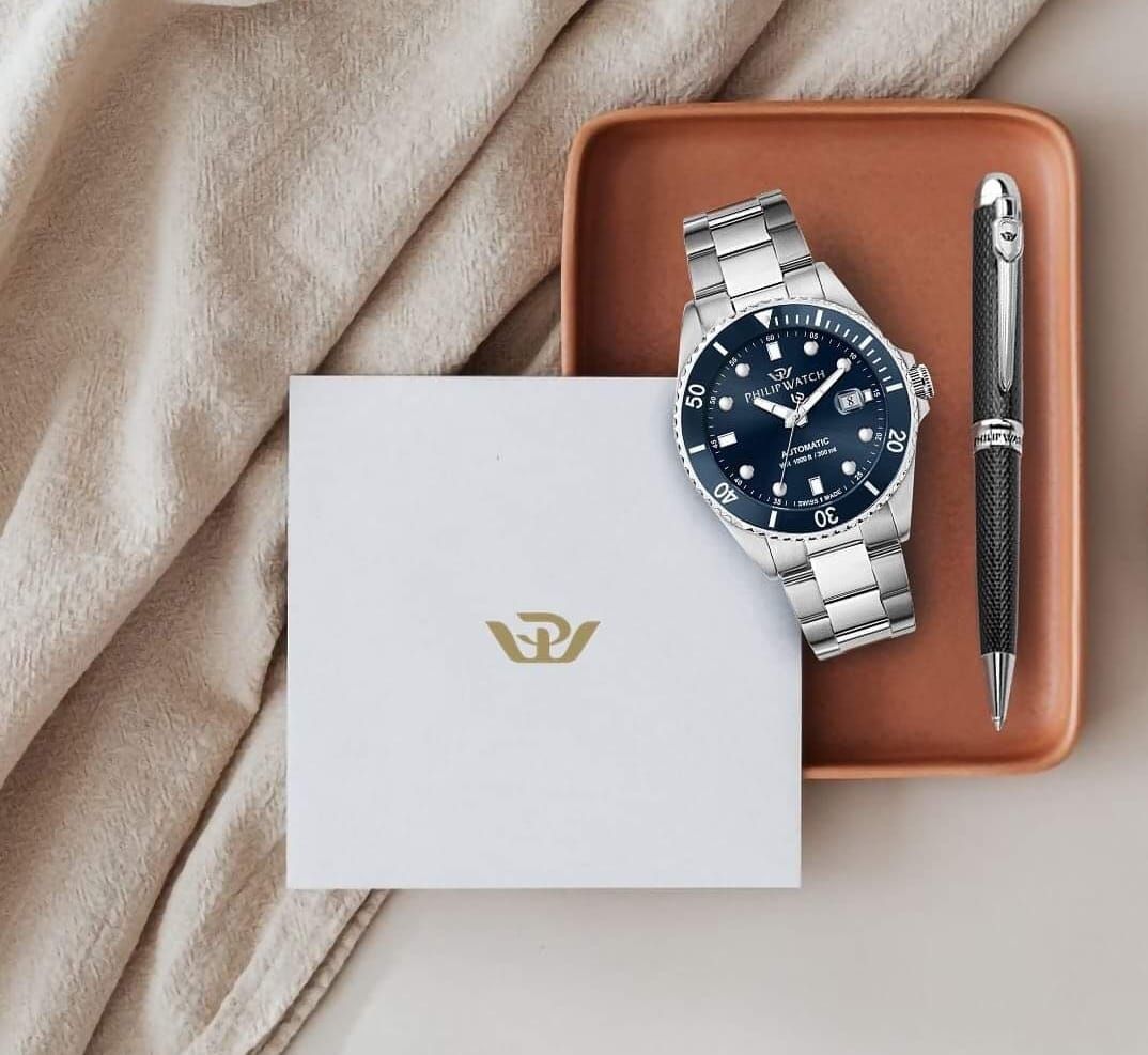 Scatola per orologio bianca con logo dorato, orologio, penna su vassoio di terracotta e panno.