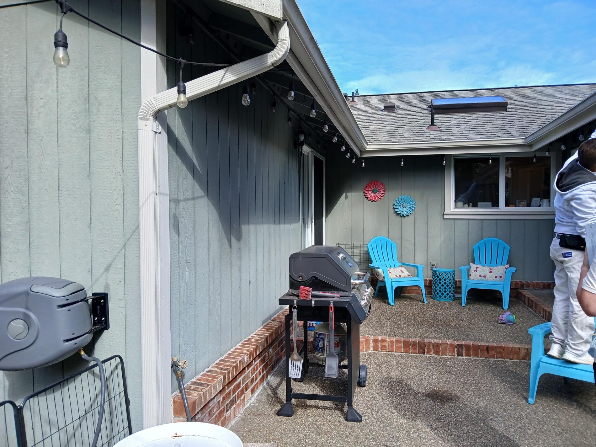 A man is standing in front of a house with a grill and chairs.