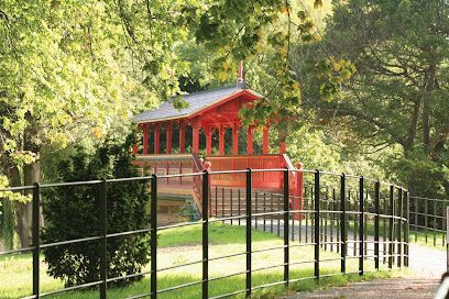 Red Asian-style gazebo behind a black metal fence on a green lawn, surrounded by trees.