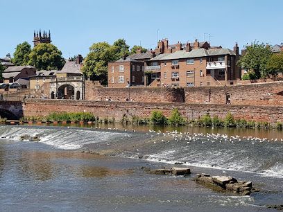 River with a weir, brick wall, and buildings in Chester, England, under a blue sky.