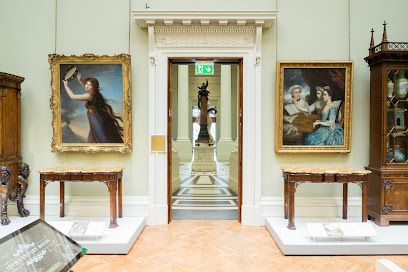 Art gallery interior with paintings flanking a doorway leading to a statue. Wooden furniture and light-colored walls.