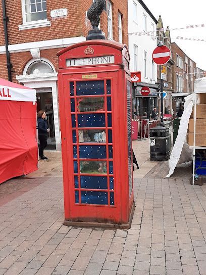 Red telephone box on a brick street with decorative interior. Person nearby, shops in the background.