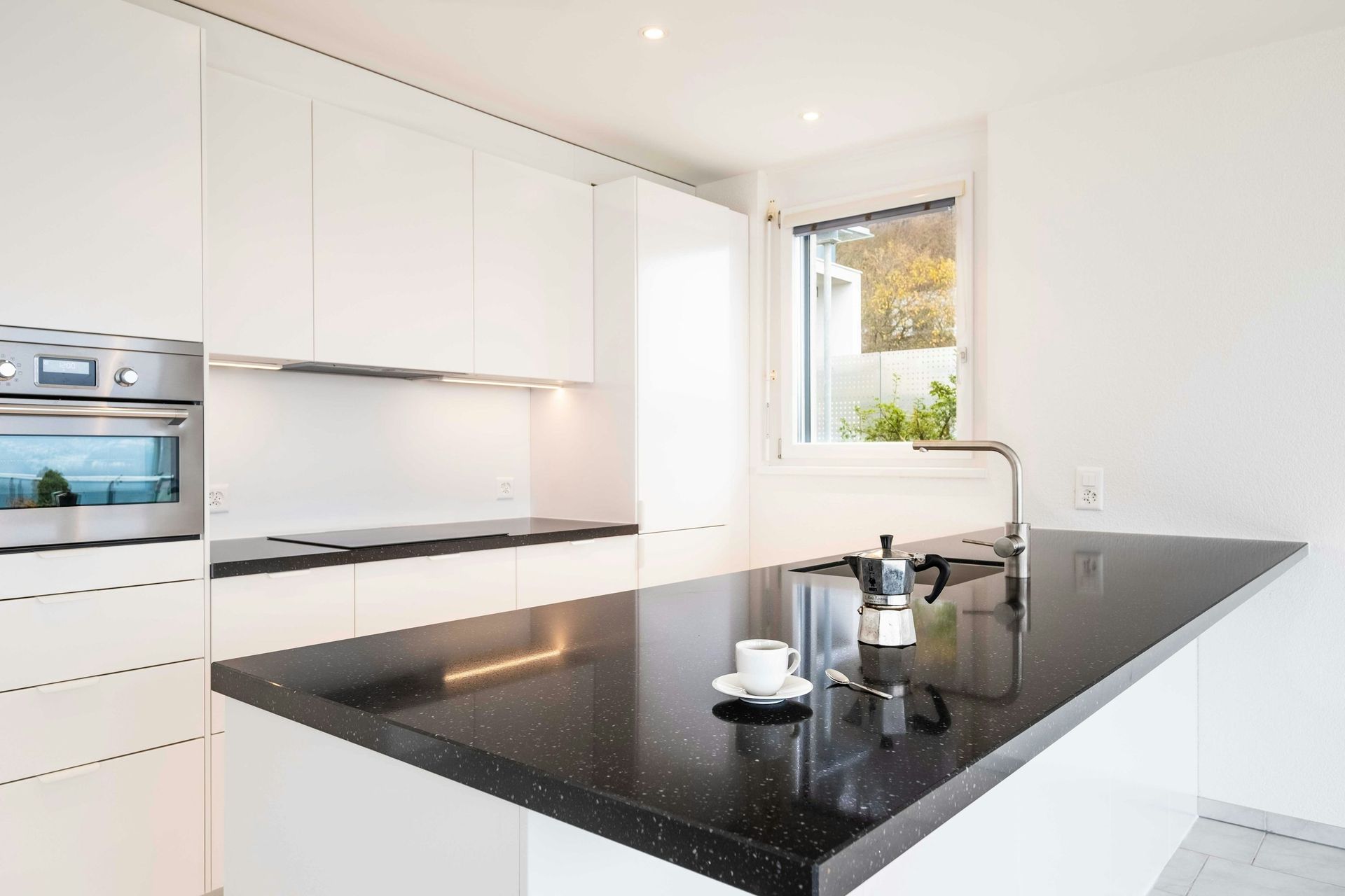 Modern white kitchen with black speckled countertop island, coffee maker, and window.