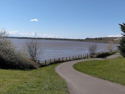 Paved path curves along a lake under a clear, blue sky. Green grass and trees frame the scene.
