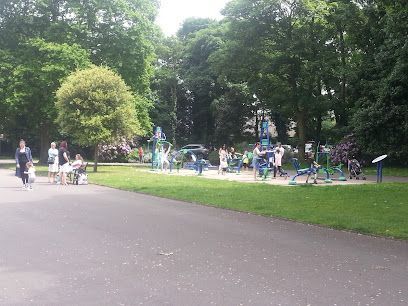 Park with outdoor gym equipment. People use the machines, some walking nearby. Trees, lawn and asphalt path.