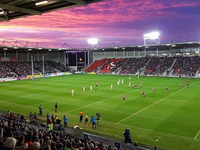 Rugby match at a stadium under a purple and pink sunset; players on the field, spectators in the stands.