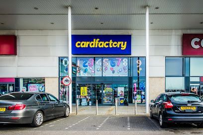 Exterior view of a Card Factory store with cars parked in front. Blue sign, beige and red building.