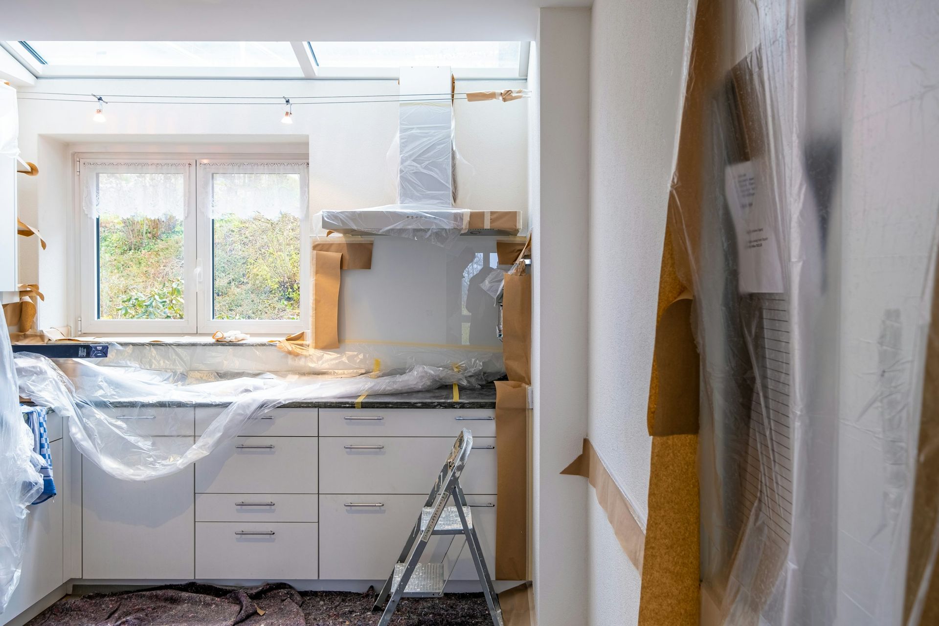Kitchen under renovation, with white cabinets, covered surfaces, and ladder. Sunlight through window.