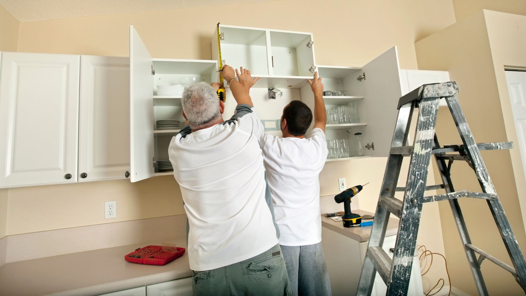 Two men installing a white kitchen cabinet using a tape measure and drill near a ladder.