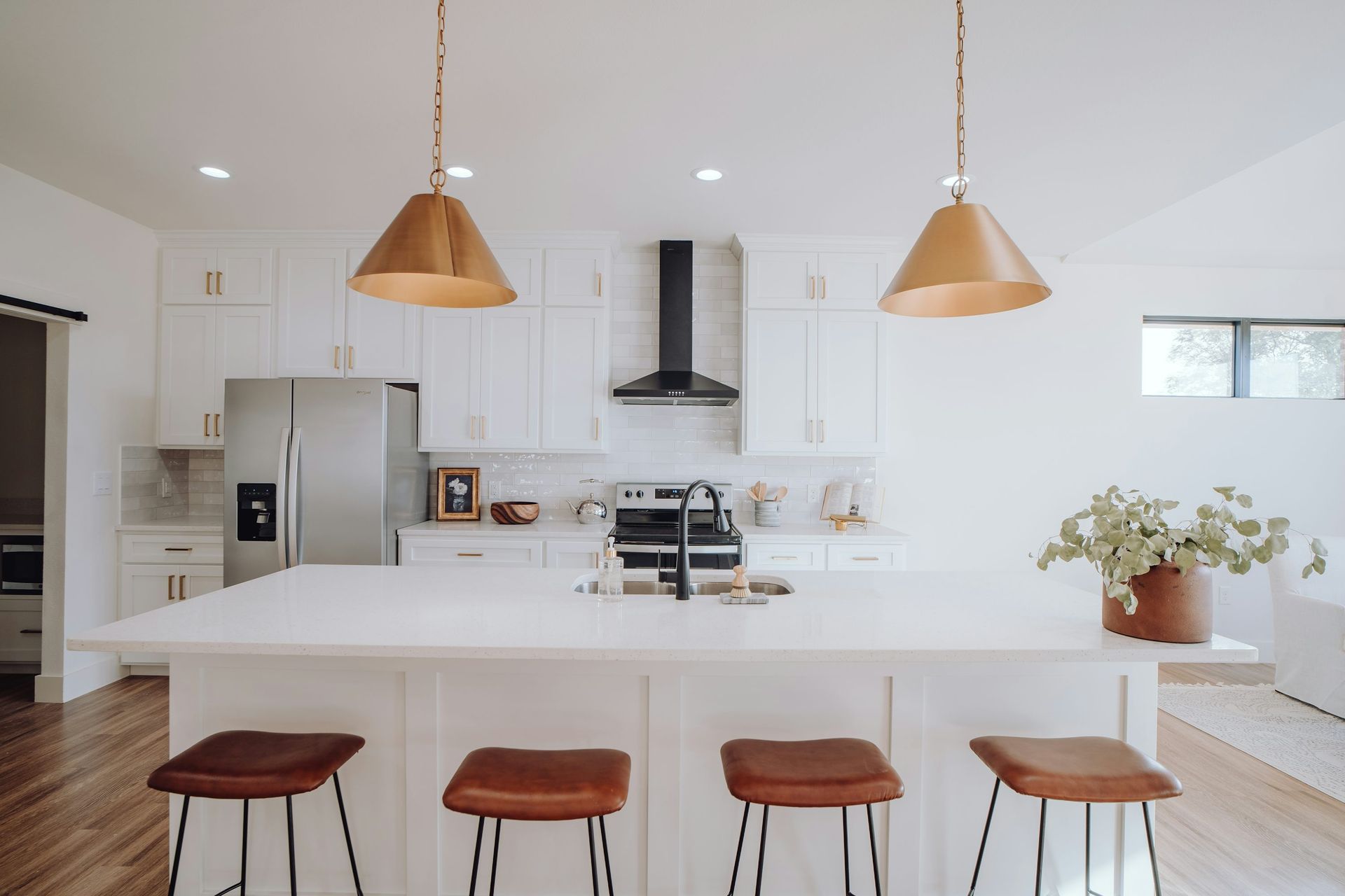 Modern white kitchen with island, bar stools, pendant lights, stainless steel appliances, and wooden floors.