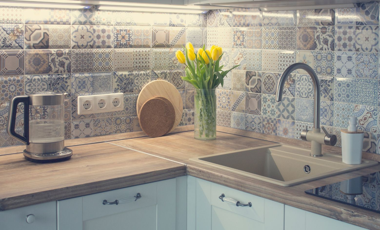 Sunlit kitchen counter with sink, kettle, vase of yellow flowers, and patterned tile backsplash
