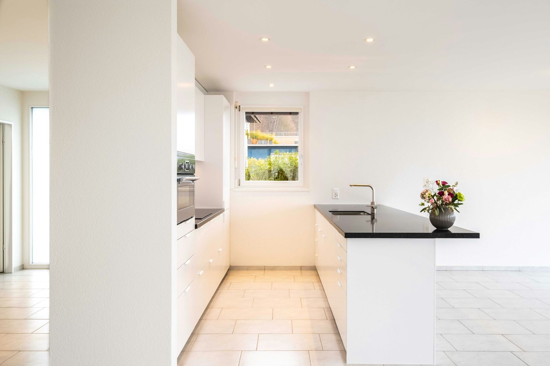 Modern white kitchen with black countertops, a window, and flowers.