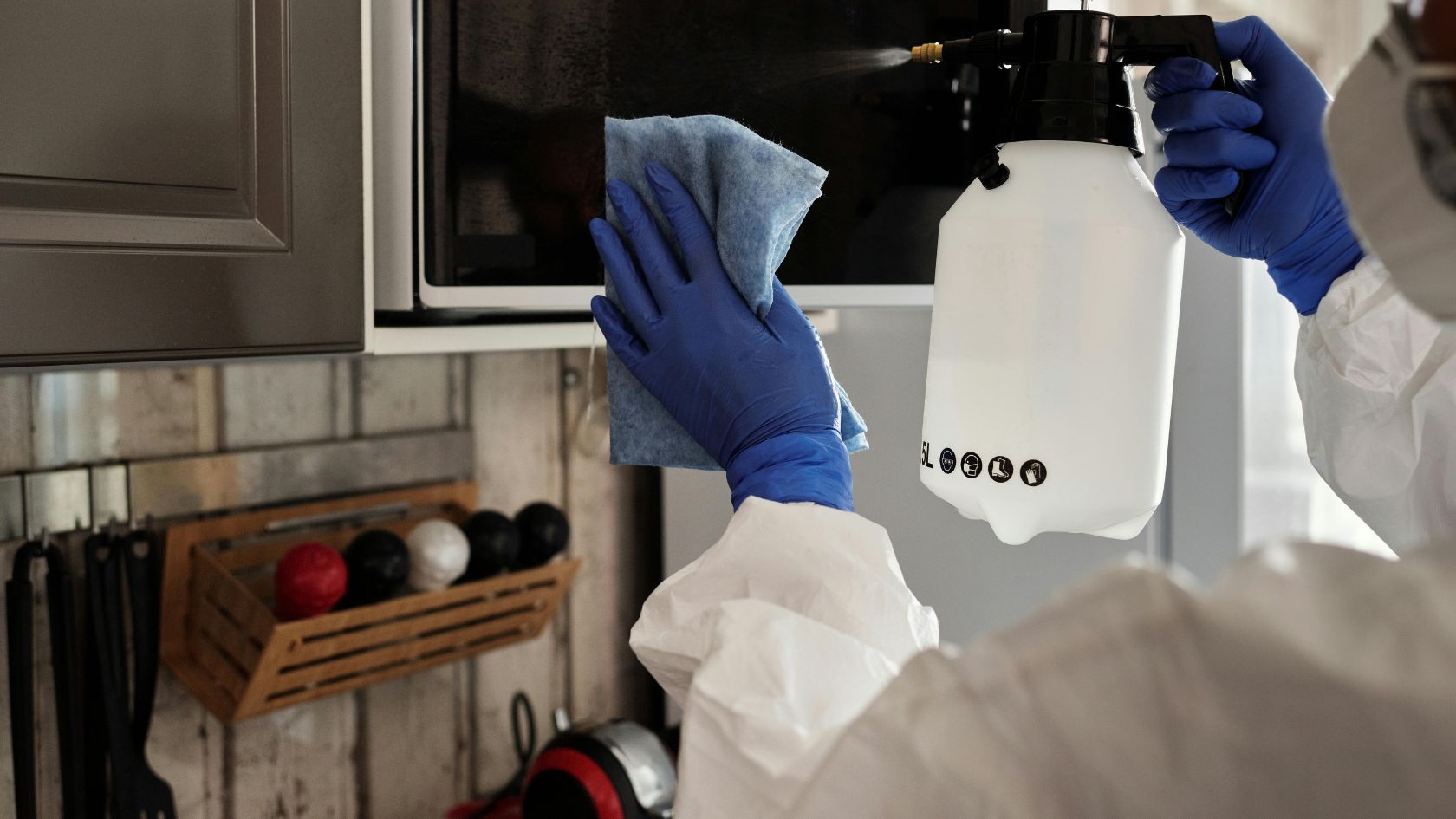 Person in protective suit sprays and wipes a microwave in a kitchen.