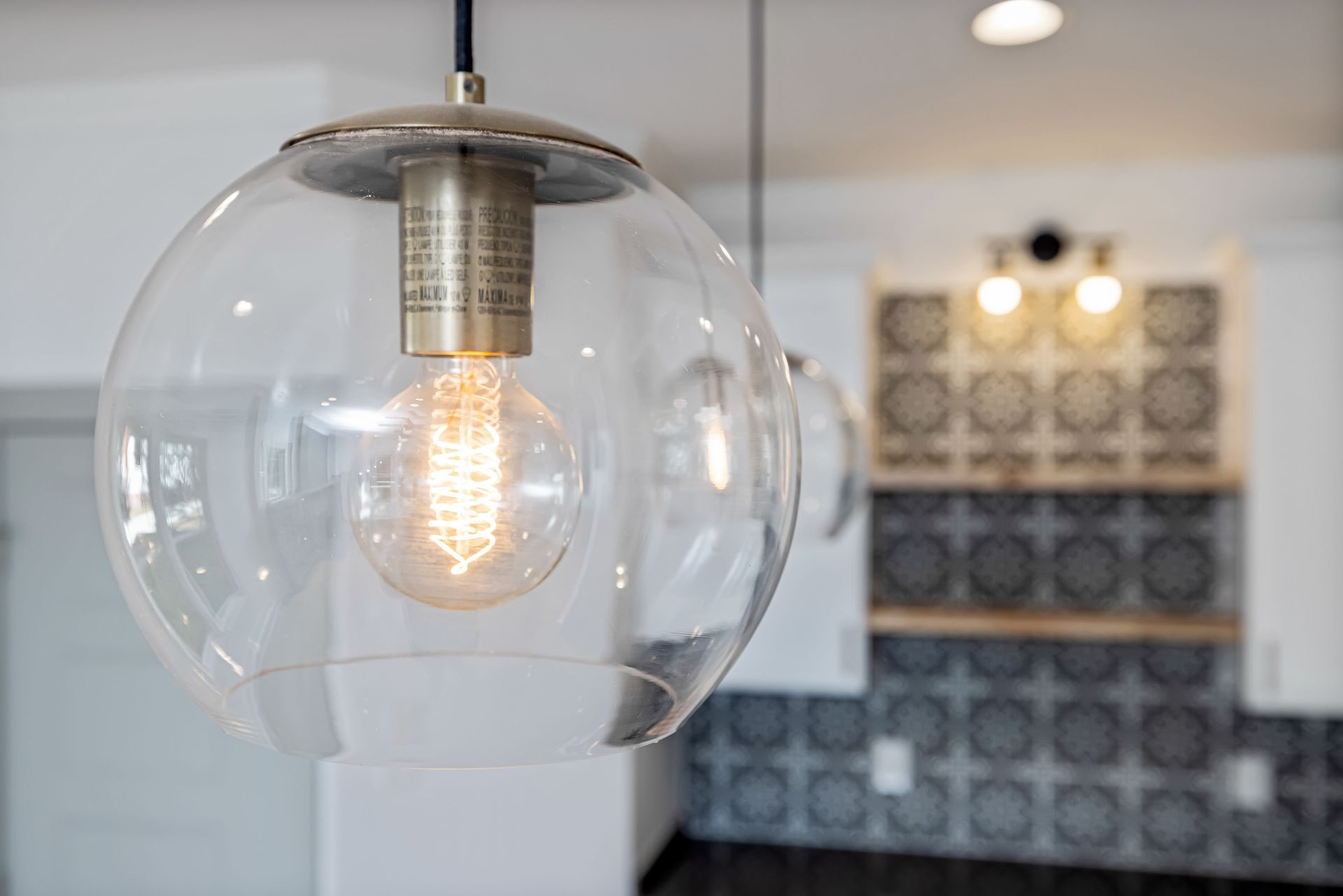 Clear glass pendant light with glowing bulb in a modern kitchen near patterned tile wall