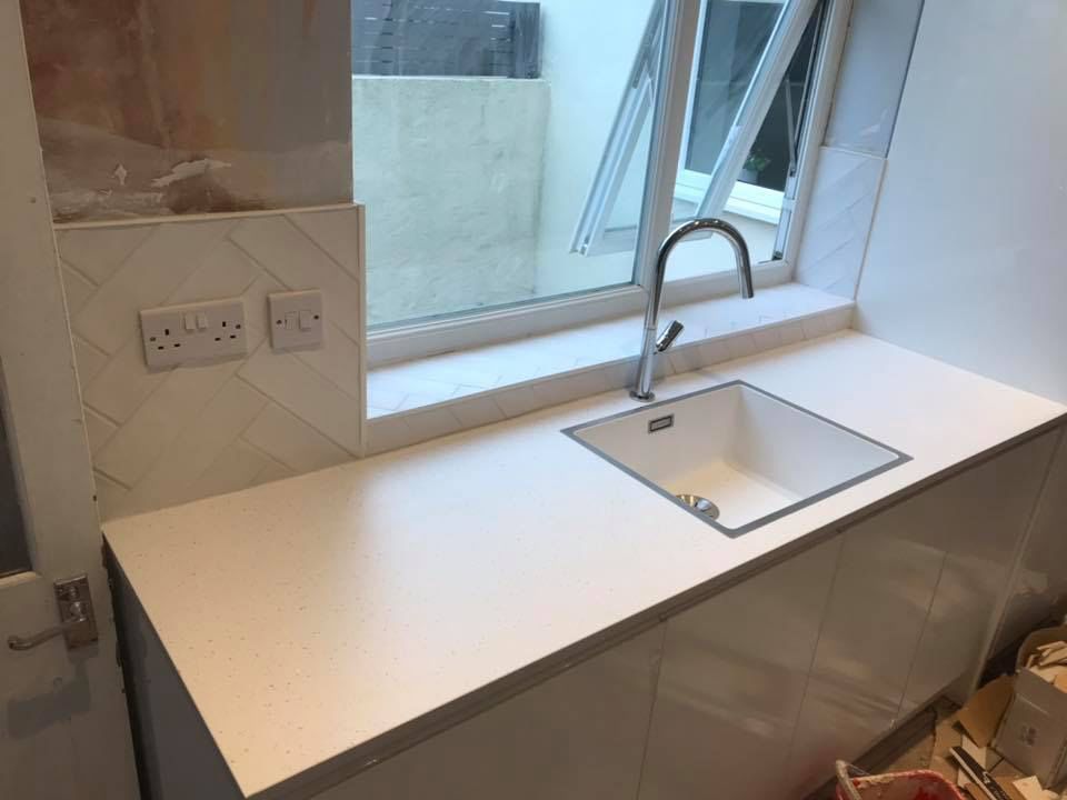 White kitchen countertop with sink and faucet near a window, with white tile backsplash and outlets.