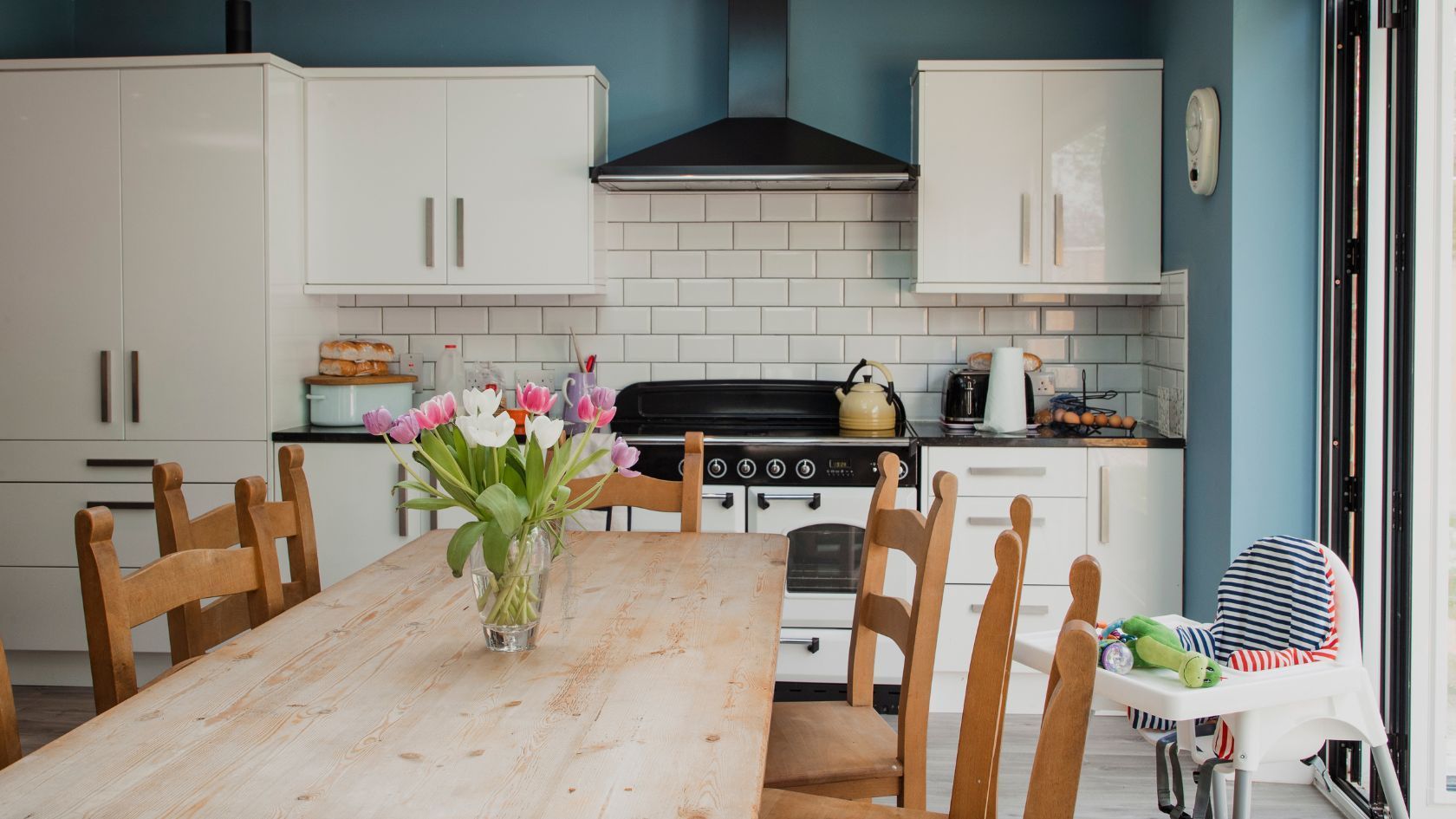 Kitchen with white cabinets, blue wall, wooden table with flowers, stove, and a high chair.