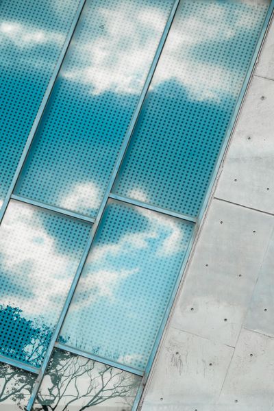 Blue perforated glass panels reflect a cloudy sky, next to a concrete wall.