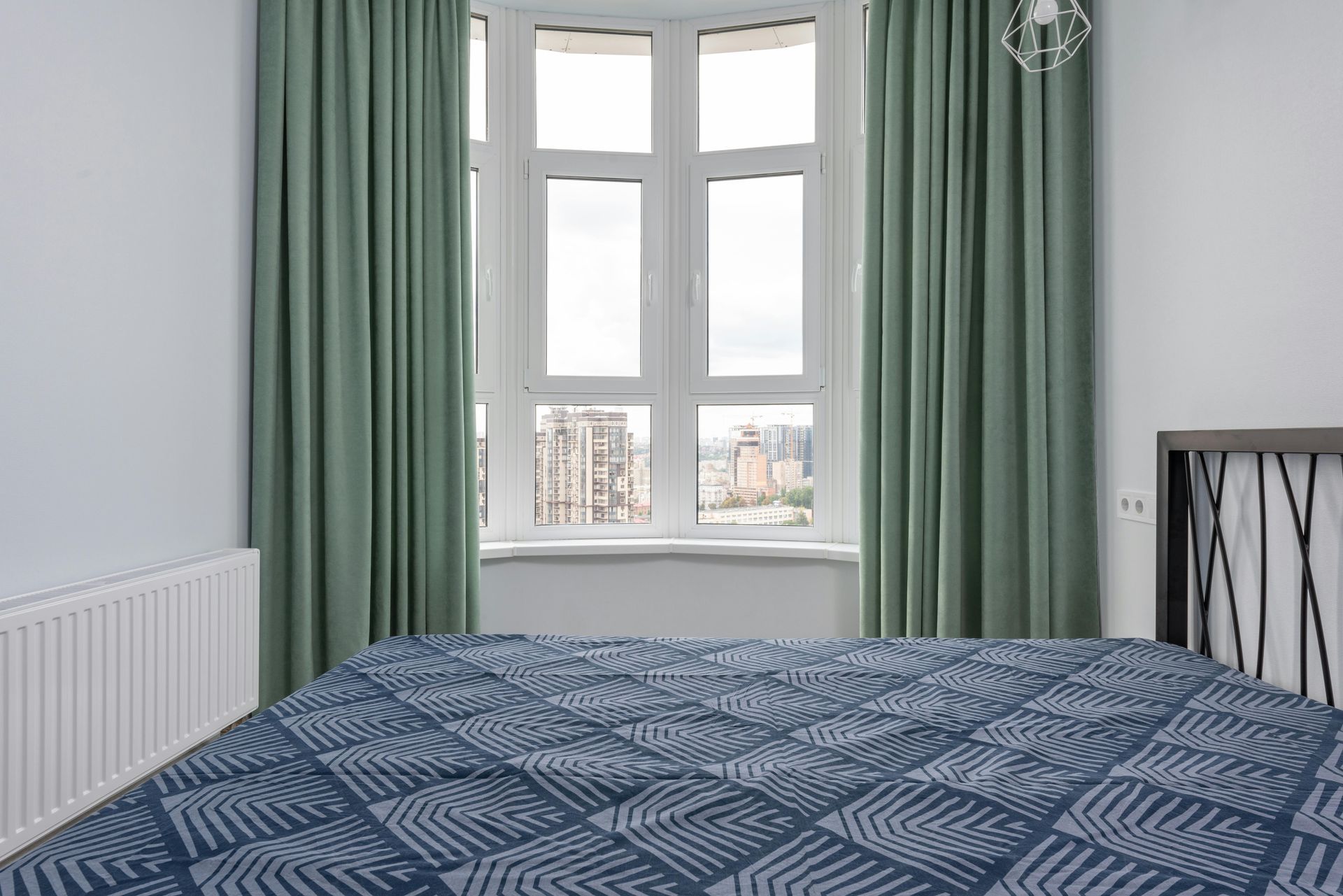 Bedroom with a bed, window, curtains, and radiator. Green curtains, blue patterned bedspread, and city view.