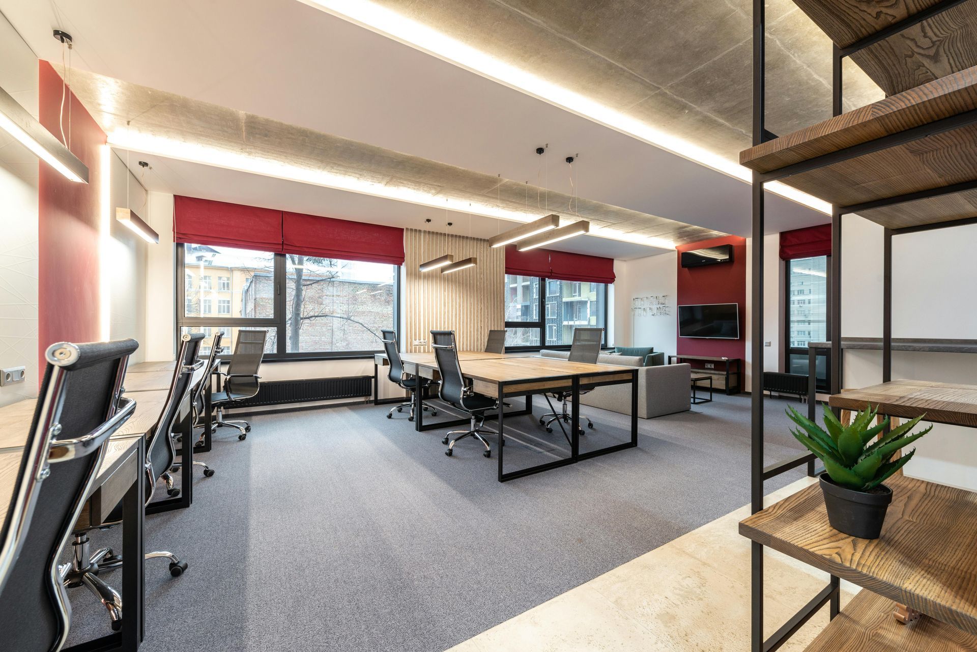 Modern office space with desks, chairs, large windows, gray carpet, and a bookshelf.