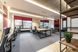 Modern office space with desks, chairs, large windows, gray carpet, and a bookshelf.