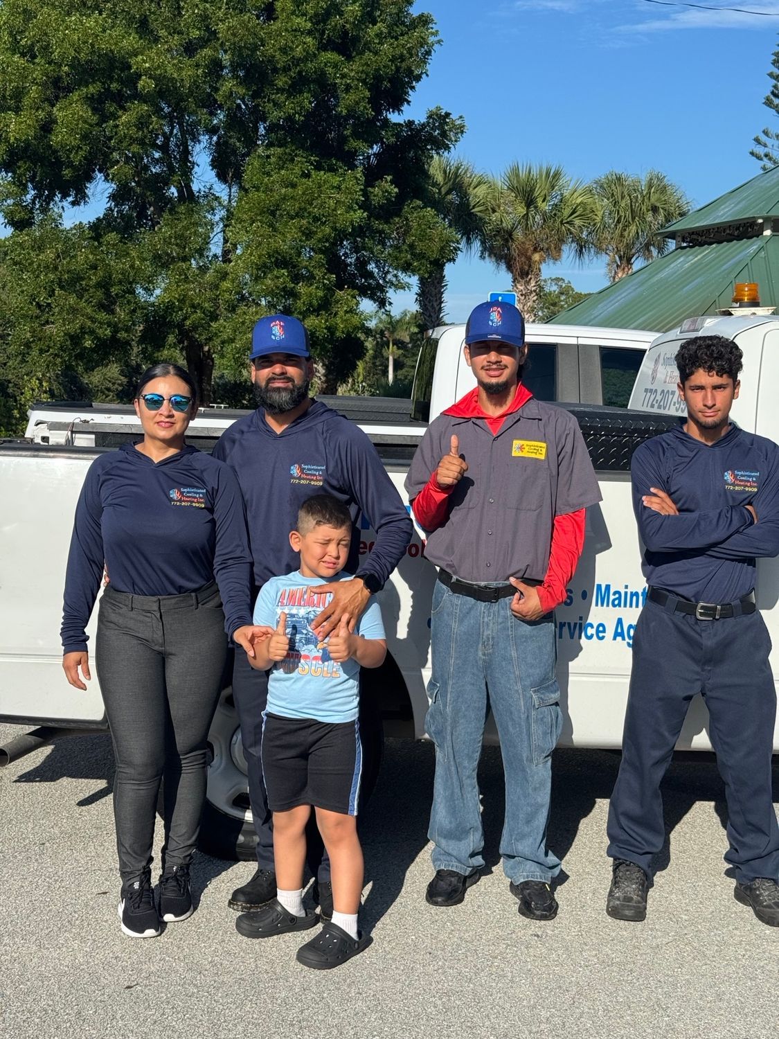 Group of five people in work attire pose by a white truck; blue tops, a young boy, thumbs up, sunny day.