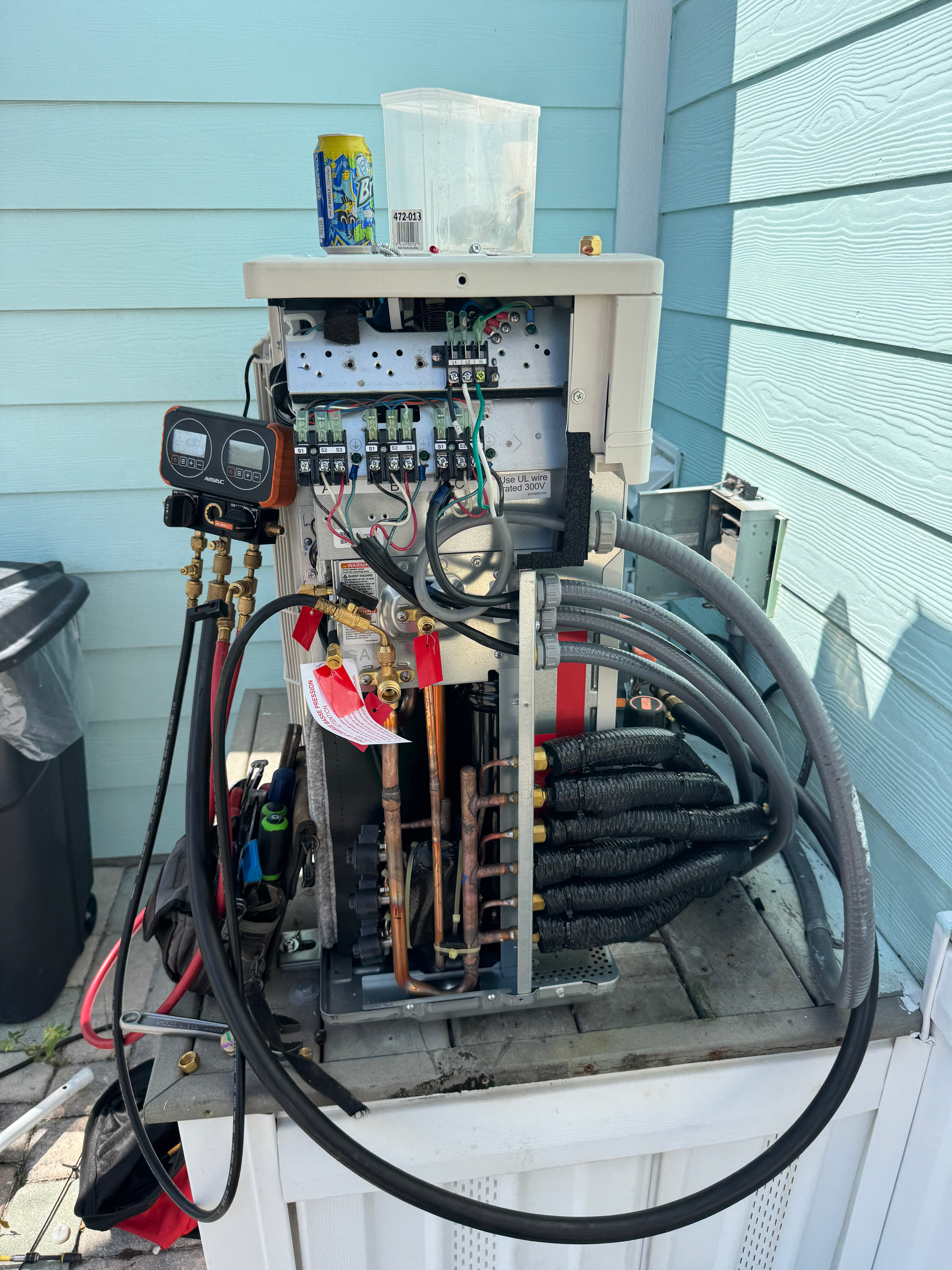 Exposed HVAC unit on a rooftop with wires, pipes, and gauges. Blue siding and trash can in the background.
