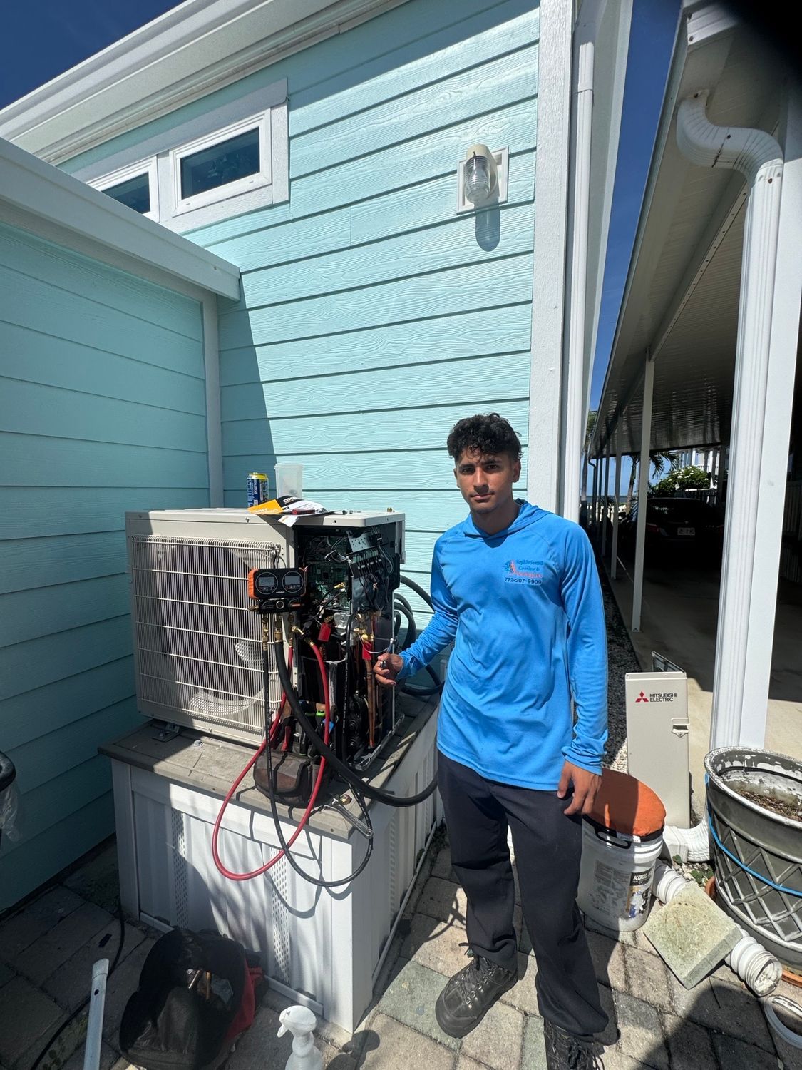 HVAC technician next to an air conditioning unit on a bright sunny day, wearing blue shirt and dark pants.