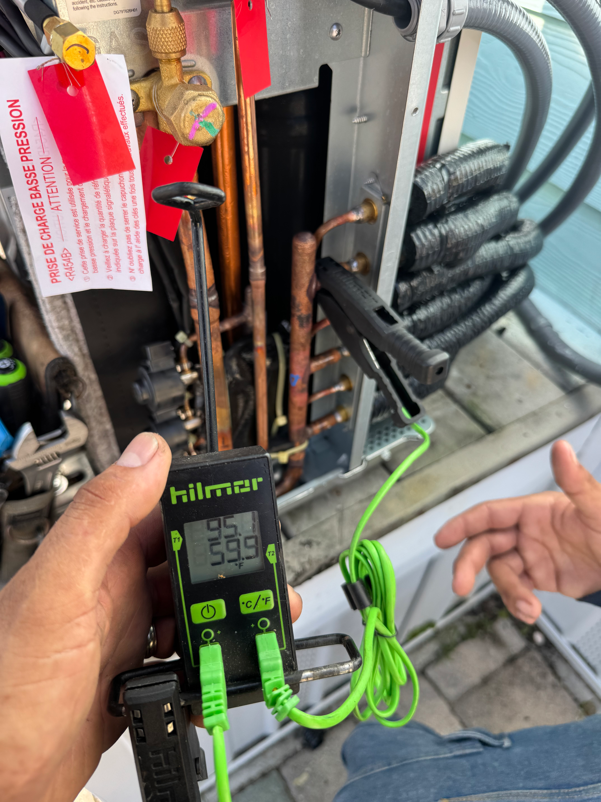 A person using a temperature reader on AC unit's copper pipes outdoors. Display reads 59.5°F.