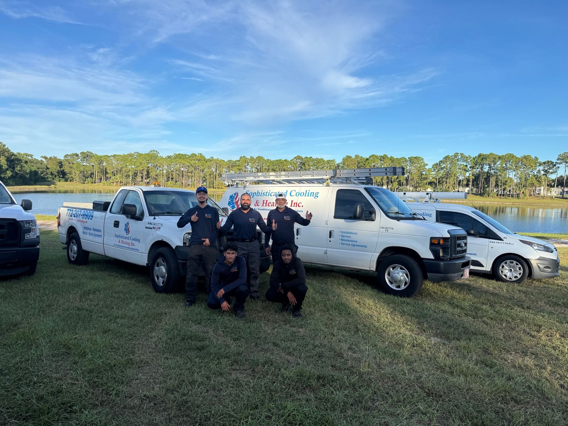 Workers in front of work vehicles near a lake, some giving thumbs up. Blue sky, green grass.