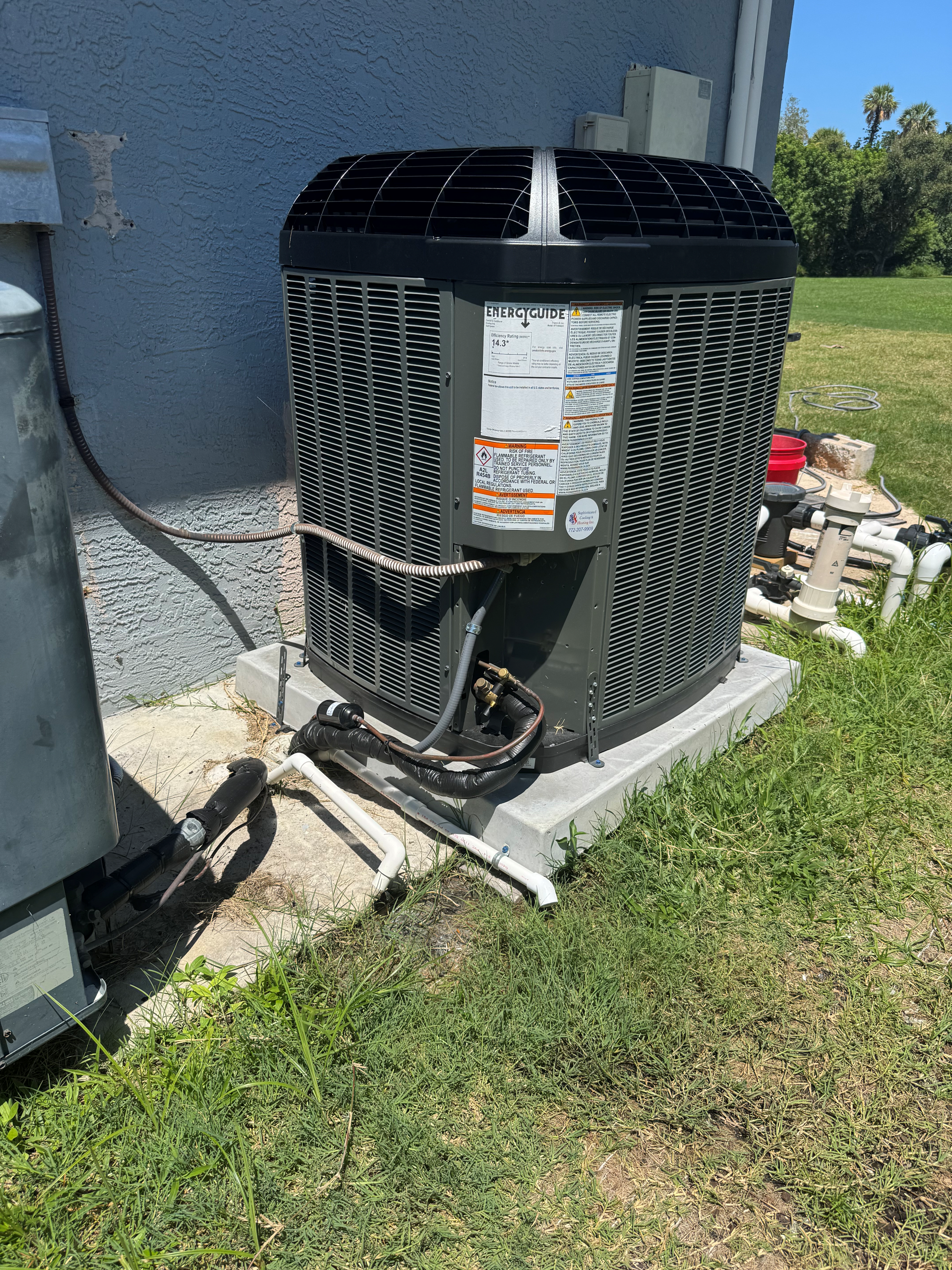 Air conditioning unit outside, next to a building, on a concrete pad, with pipes and wires visible.