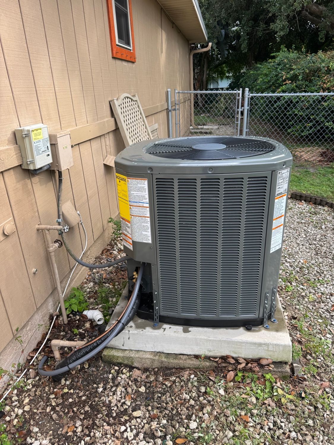Air conditioning unit on concrete slab next to a house with a chain-link fence in the background.