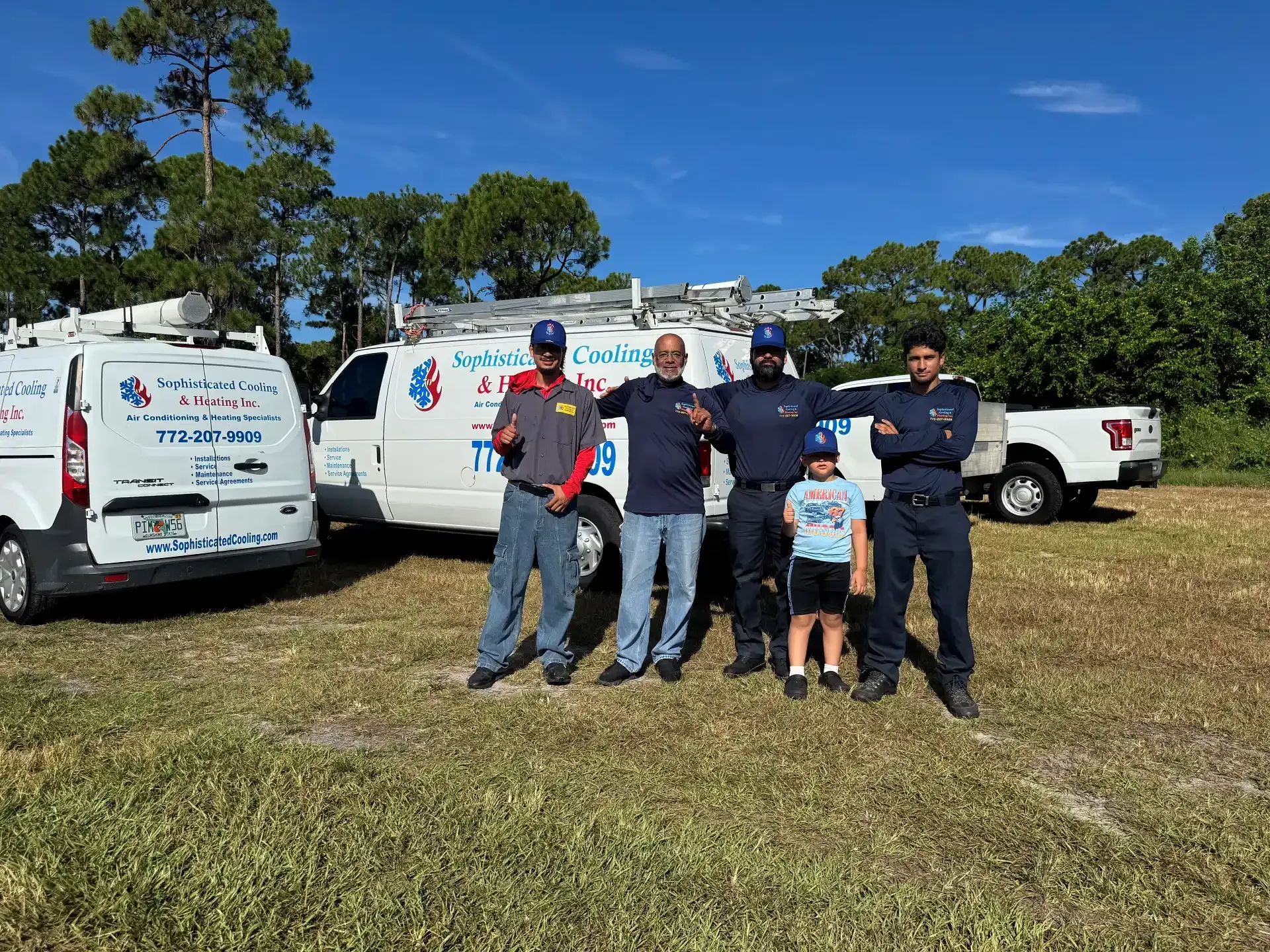 HVAC crew posing by service vans on a sunny day. The men are wearing work uniforms and a young child is present.