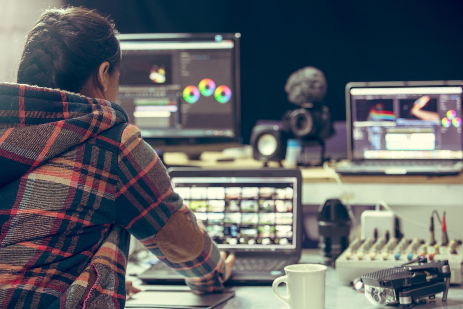 Person editing video on multiple screens, with coffee cup, microphone, and speakers.