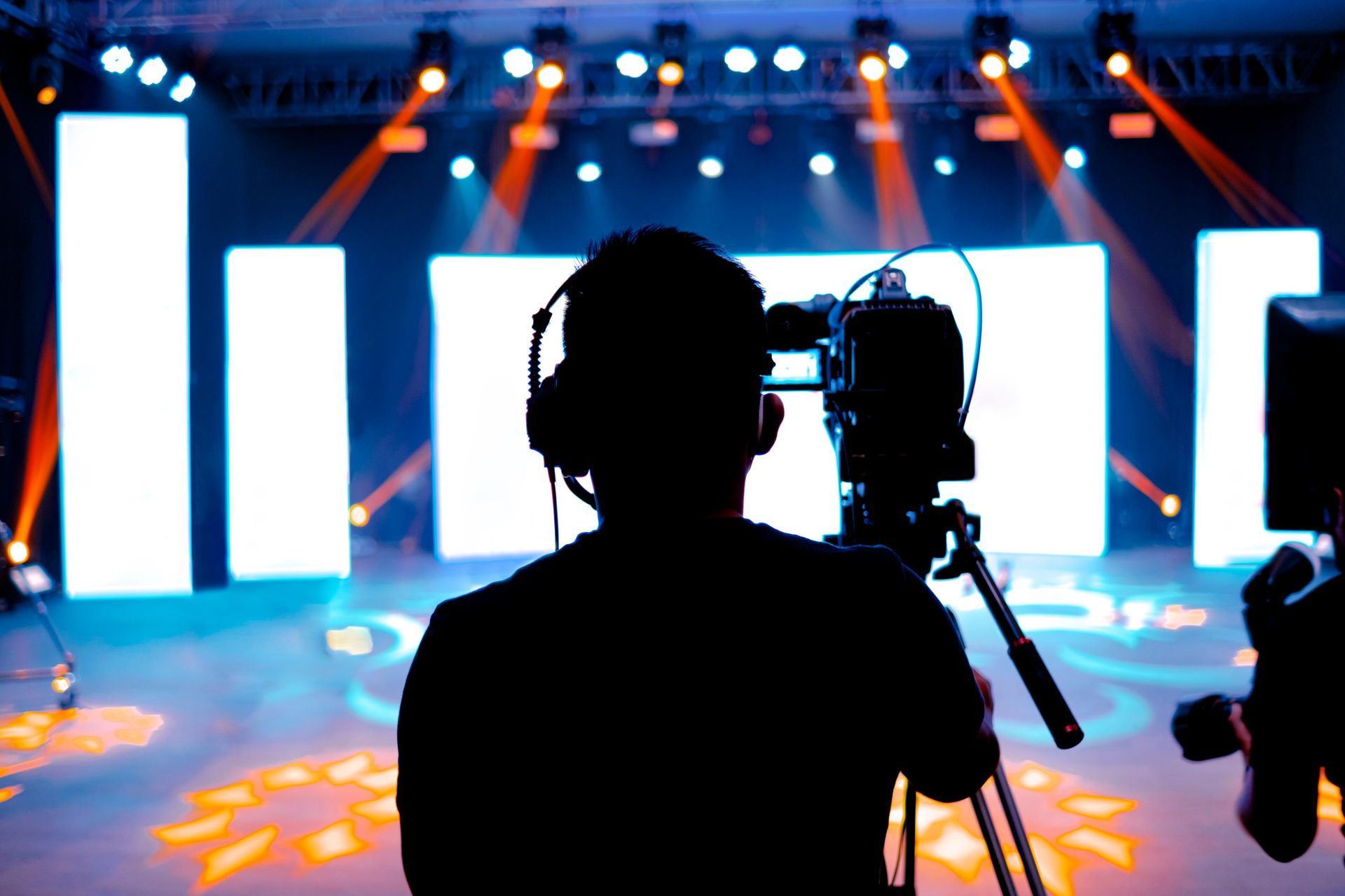 Silhouette of a person with headphones operating a camera on a tripod, stage with bright lights in background.