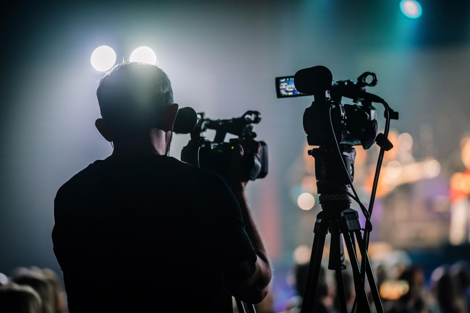 Person filming a concert with two cameras on tripods, stage in background.
