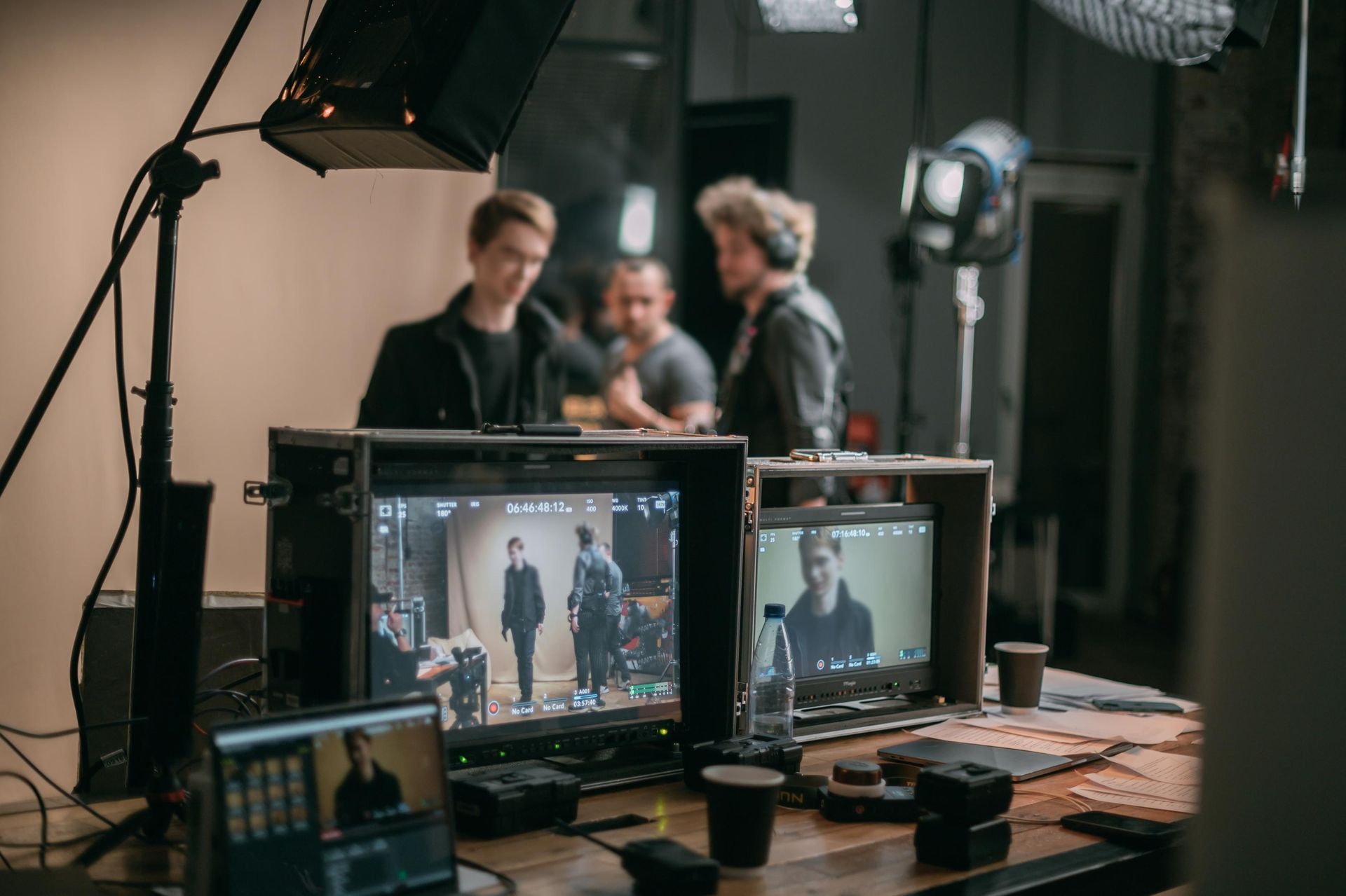 Film set: Monitors show actors, crew in the background, studio lights, and equipment on a table.