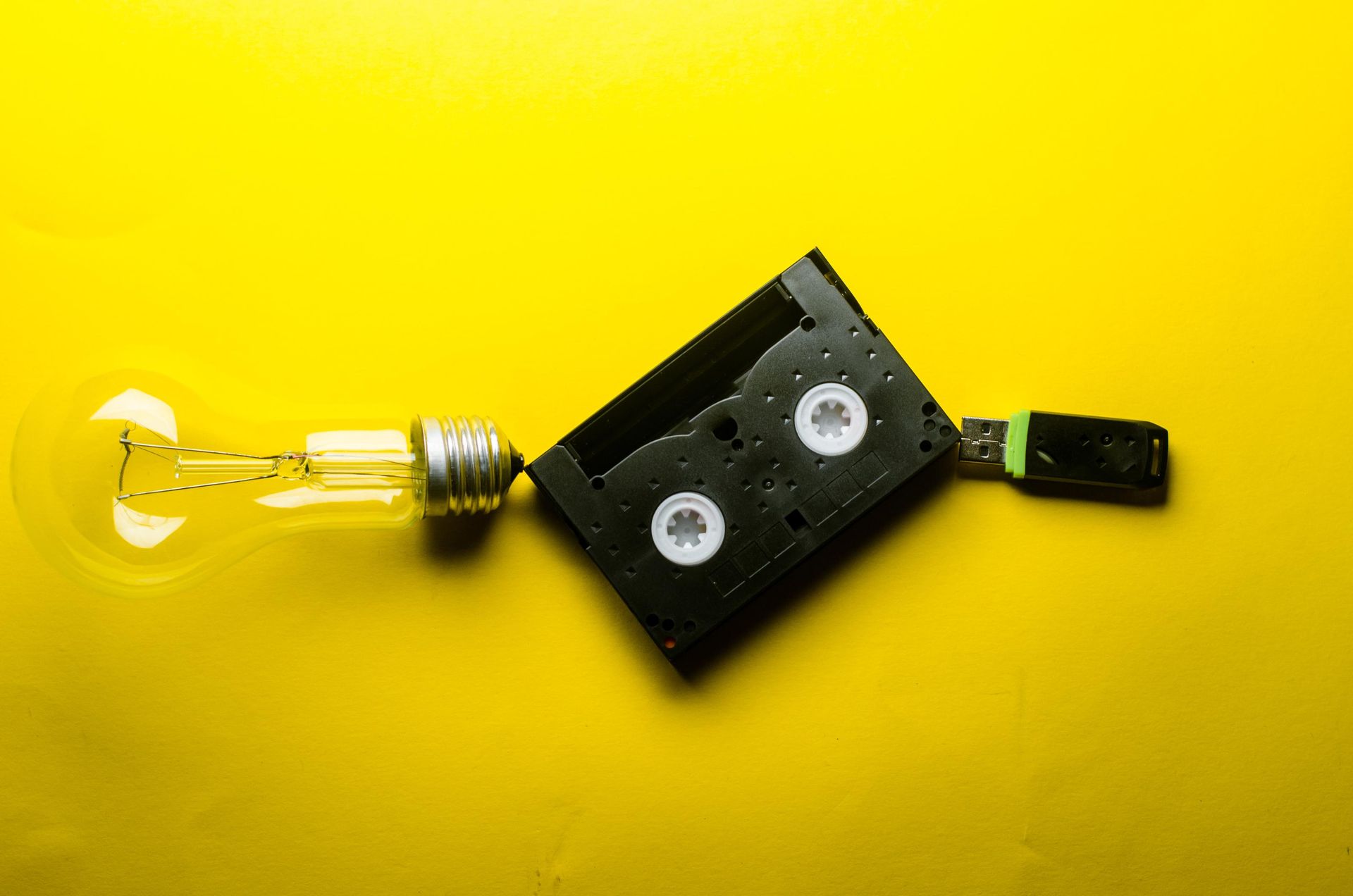 Lightbulb connected to a VHS tape and a USB drive on a yellow background.