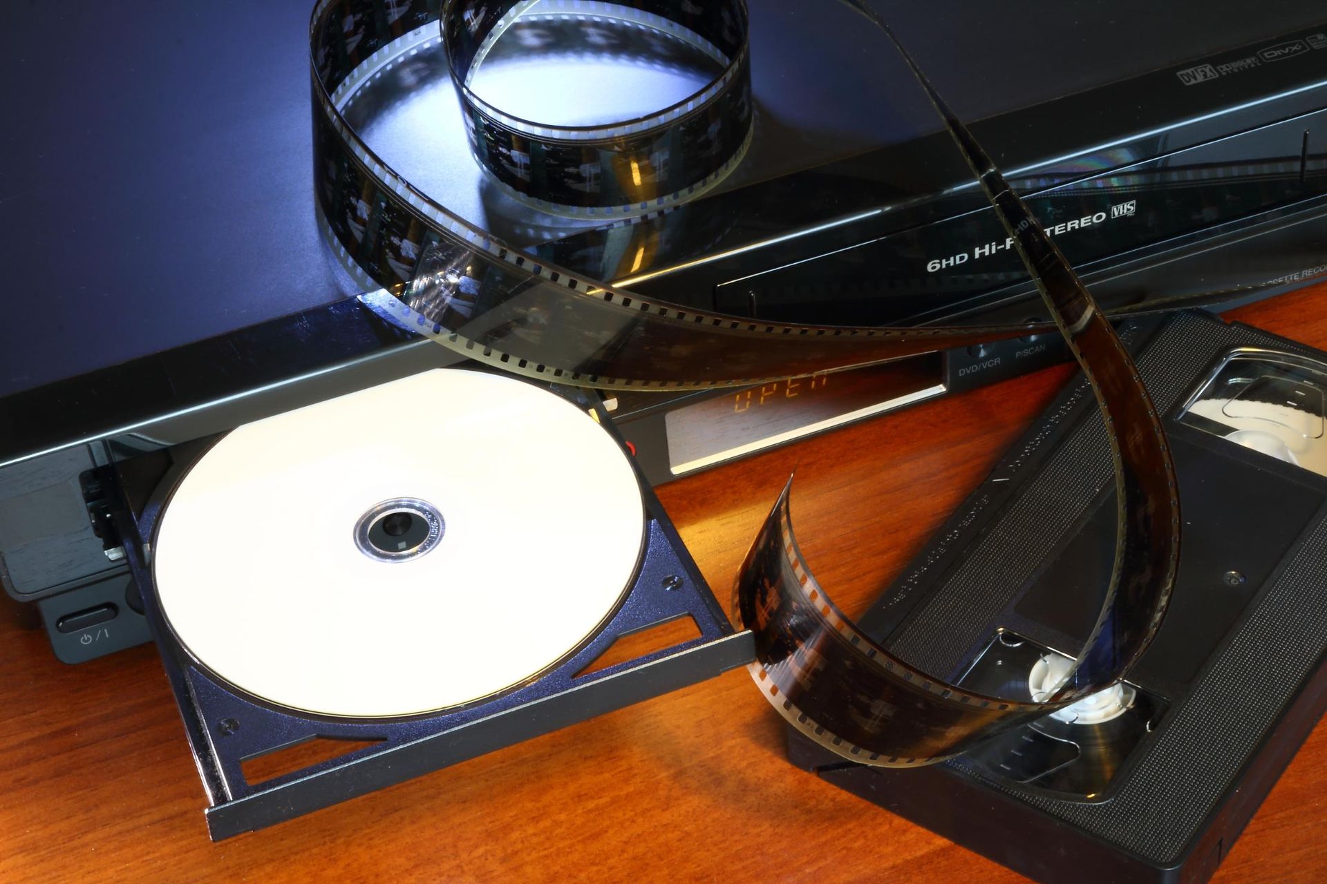 DVD player with open tray holding a disc, film strip, and a VHS tape on a wooden surface.