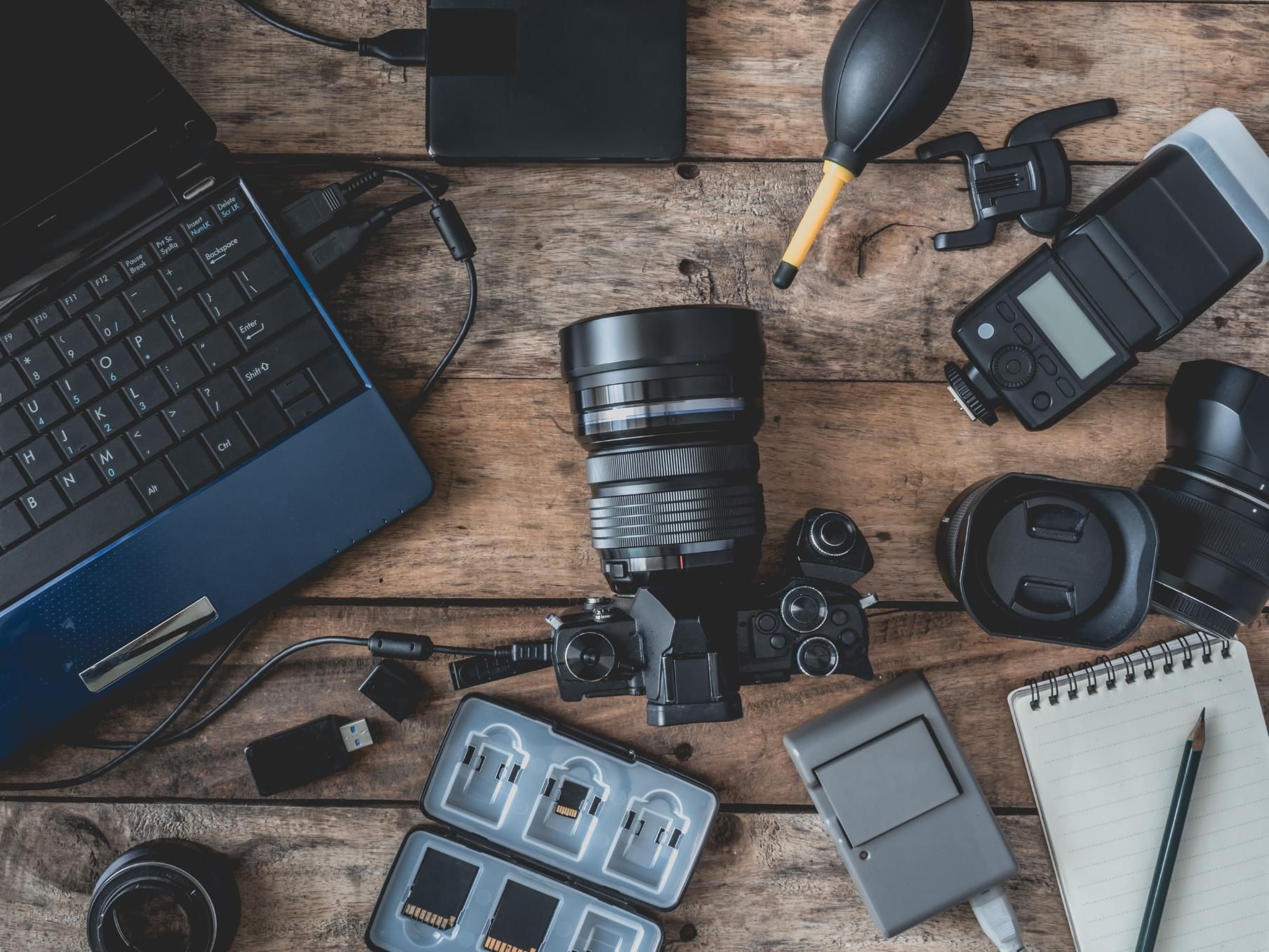 Photographer's tools on wooden table: laptop, camera, lenses, flash, SD cards, notebook, and cleaning tools.