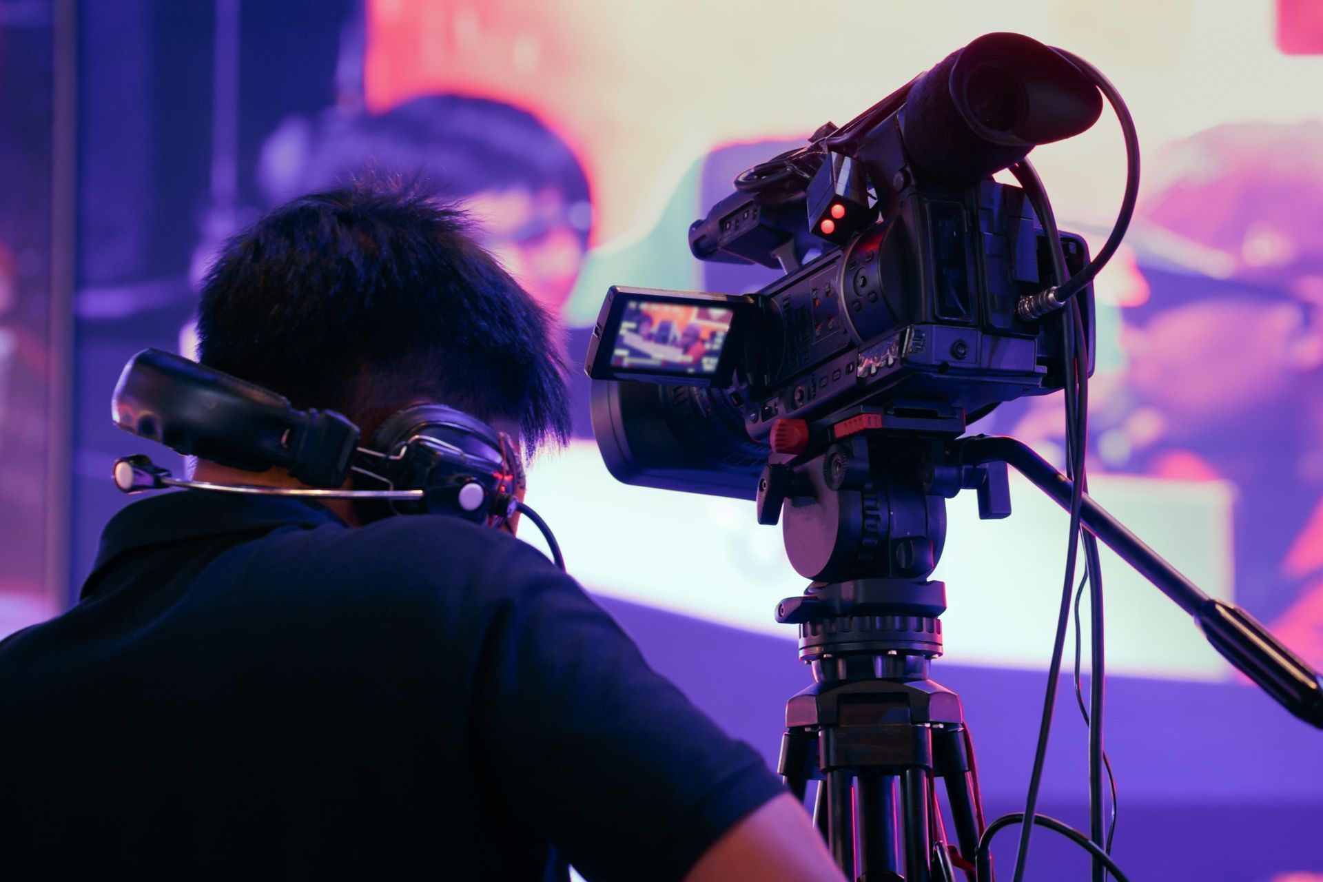 Person with headphones operates a video camera on a tripod, filming an event with a large screen in the background.