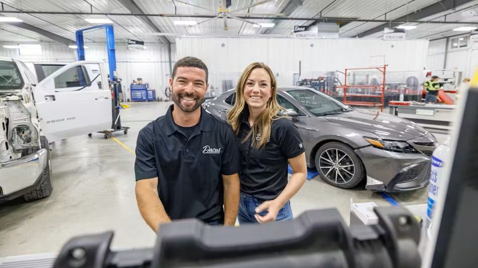 Owners smiling in auto body shop, gray car and truck visible, shop interior.