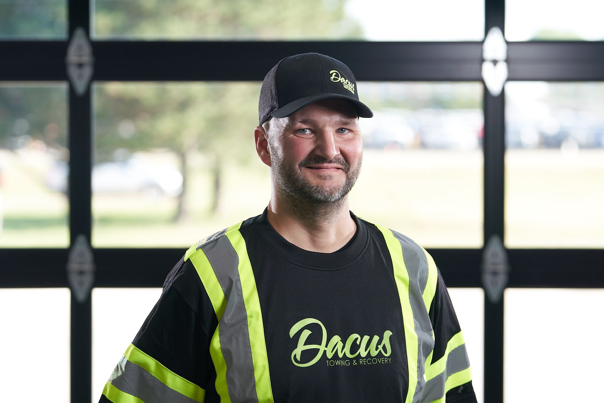 Man in reflective vest and cap with company logo smiles in front of a window.