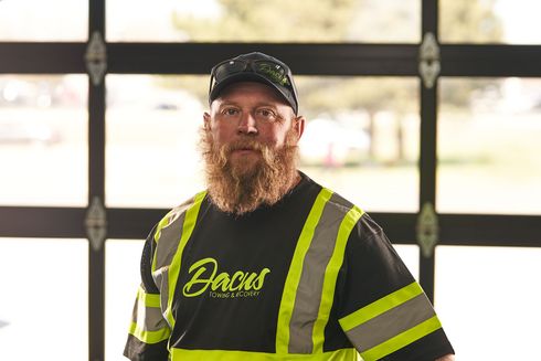 Man with red beard, wearing a black shirt with reflective stripes and a cap, standing in front of windows.