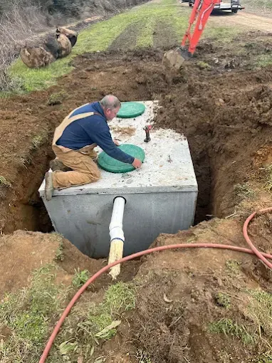 A worker kneels on a concrete septic tank installed in a muddy trench, near an excavator on a grassy field.